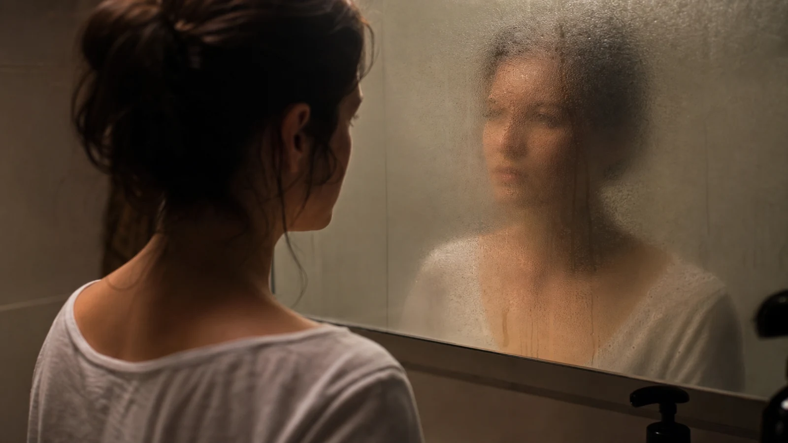 Femme de dos face à un miroir embué, reflet flou de son visage, lumière douce