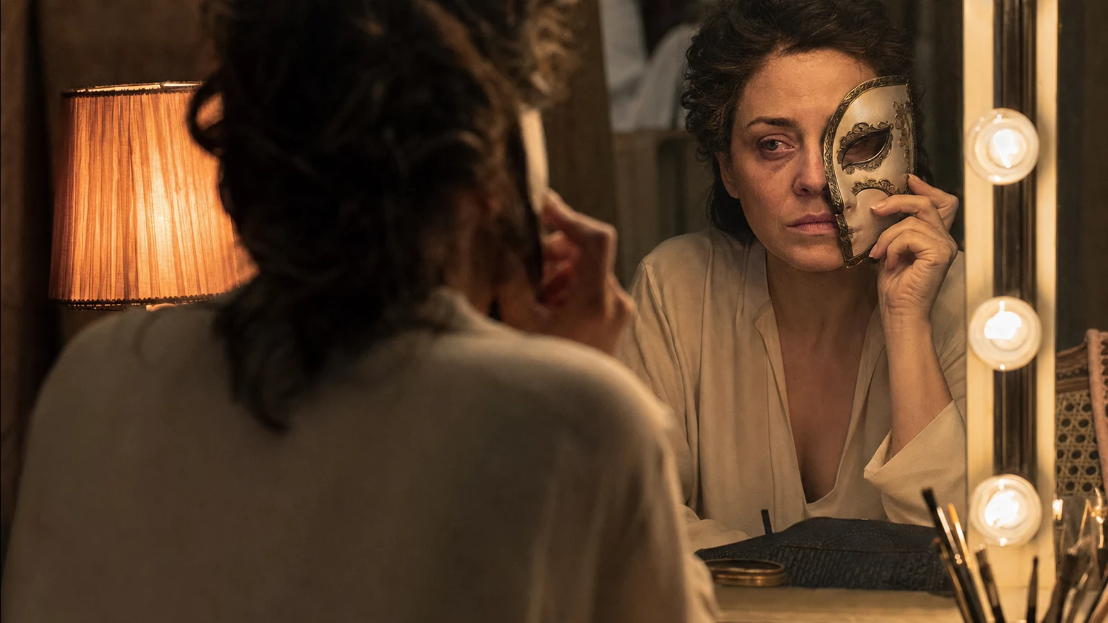 Femme en chemise claire, vue de dos devant un miroir de loge éclairé, tenant un demi-masque clair orné contre son visage reflété