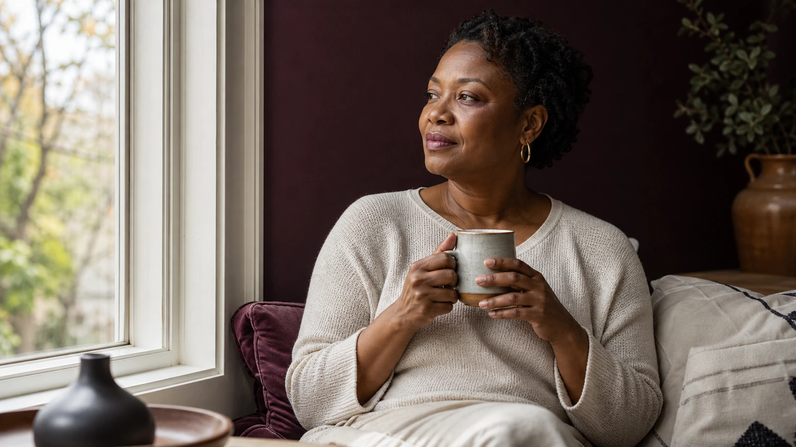 Femme noire d’environ 48 ans assise près d’une fenêtre, tasse entre les mains, lumière douce