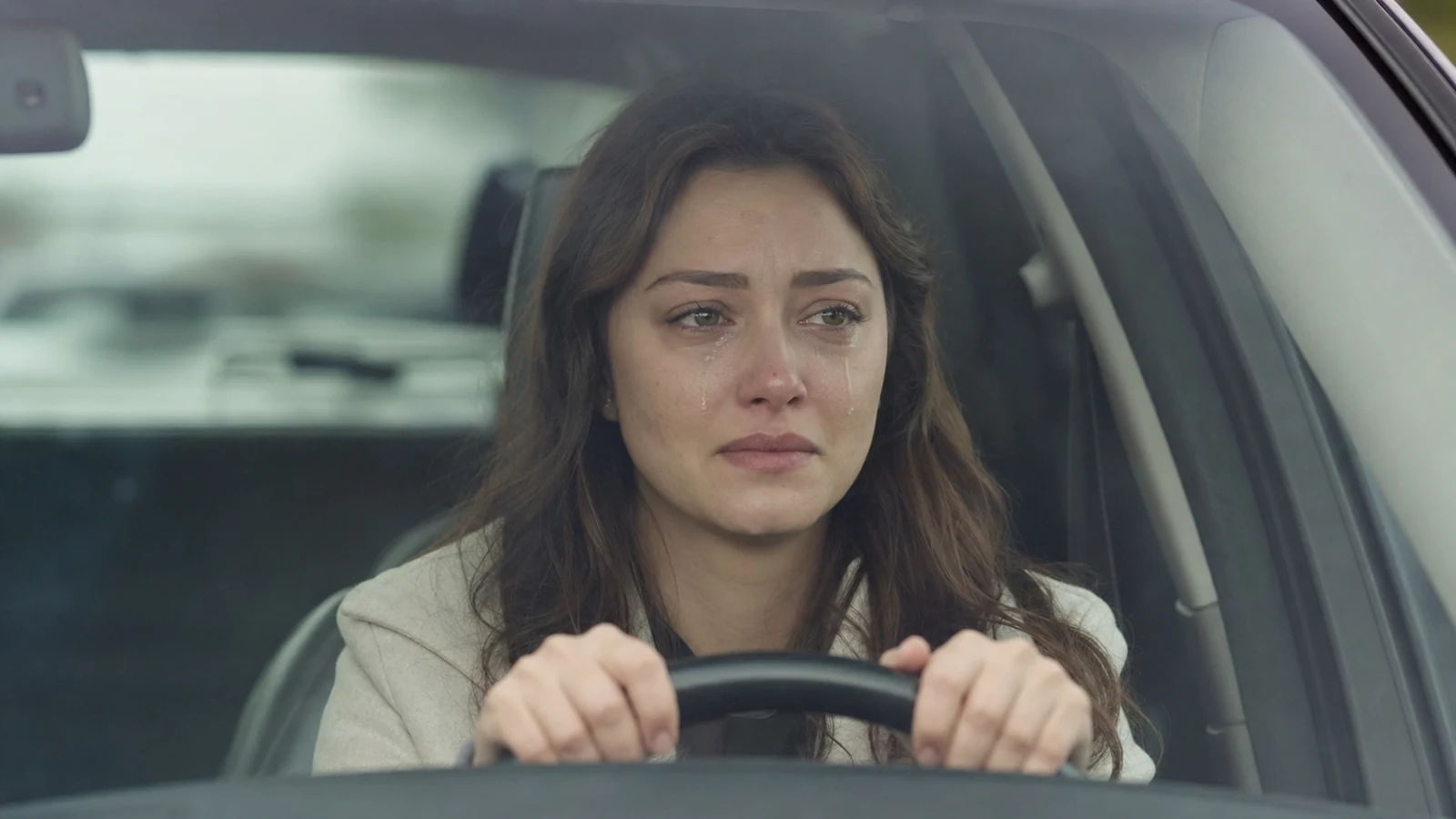 Femme aux cheveux bruns au volant d'une voiture, larmes sur le visage, manteau clair, regard vers l'avant